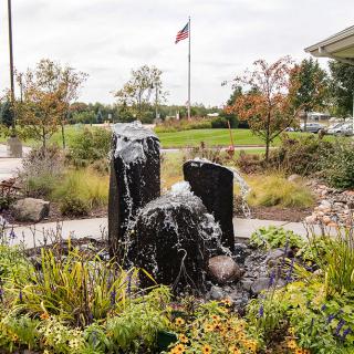 A tranquil garden scene featuring a stone fountain surrounded by lush greenery and an American flag in the background.