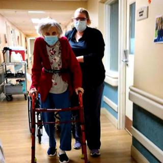A caring healthcare worker assists an elderly woman using a walker in a hospital corridor.