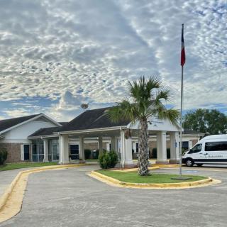 A building with a van parked in the driveway, featuring a flagpole and a palm tree under a cloudy sky.