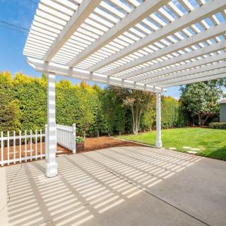A sunny backyard patio with a white pergola casting shadows on the pavement.