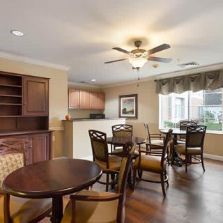 A cozy dining area with wooden tables, chairs, and a ceiling fan, adjacent to a small kitchenette and a large window with city views.