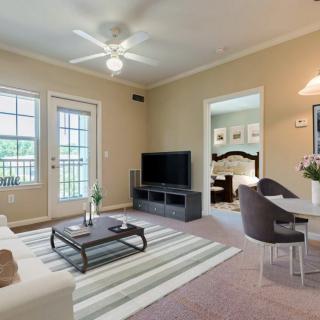 A cozy living room with a TV, sofa, armchair, and coffee table, featuring neutral tones and natural light from large windows.