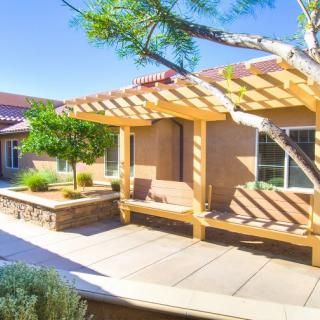 A sunny patio with a wooden pergola providing shade, surrounded by landscaping.