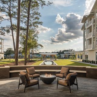 A modern outdoor seating area featuring a fire pit, surrounded by lush greenery and contemporary buildings under a cloudy sky.