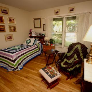 A cozy bedroom featuring a single bed with a striped blanket, a comfortable chair, and natural light streaming through the window.