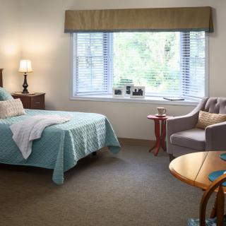 A cozy bedroom with a bed, lamp, and chair by a large window.