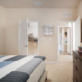 A neatly arranged bedroom with neutral tones, featuring a bed and a view through an open door into a well-lit hallway.