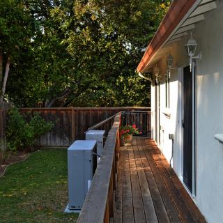 A wooden deck alongside a house with a yard and fence, illuminated by sunlight and surrounded by trees.