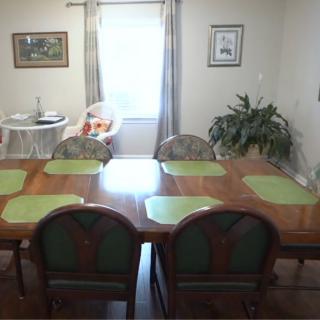 A dining room with a large table and green placemats, surrounded by chairs. A window lets in natural light, and there is artwork on the walls.