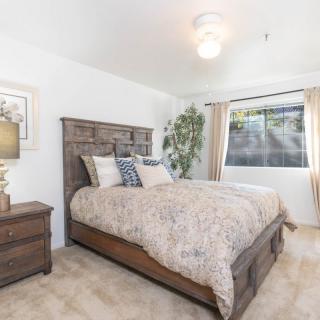 A cozy bedroom with a wooden bed frame, plush bedding, side table with a lamp, and a large window with curtains.