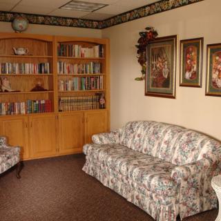 A cozy living room with a floral sofa, an armchair, a wooden bookshelf filled with books, and paintings on the wall.