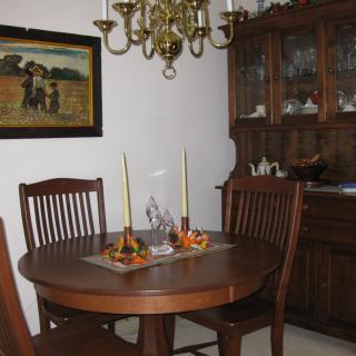 A cozy dining room with a wooden round table set for two, featuring a chandelier and a painting on the wall.