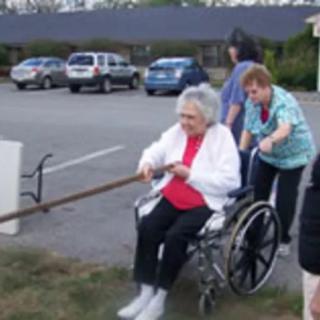 An elderly person in a wheelchair being assisted outdoors by caregivers.