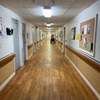 A long, well-lit hospital hallway featuring wooden flooring and walls adorned with various papers and notices.