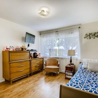A cozy bedroom with wooden flooring, featuring a bed, dresser, armchair, and a wall-mounted television.