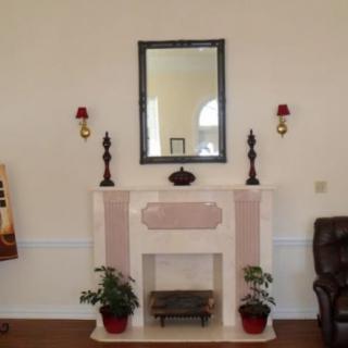 A living room with a decorative fireplace, featuring a mirror above it, flanked by two candle holders and potted plants on either side.