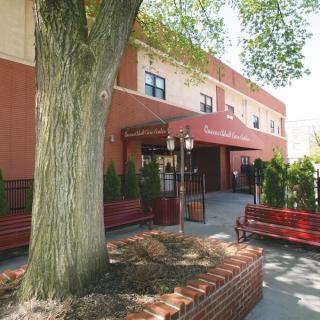 The image shows an entrance to a building with an awning, surrounded by trees and benches.