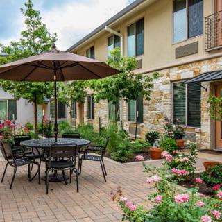 A charming courtyard with outdoor seating, featuring a table with an umbrella surrounded by lush greenery and blooming flowers.