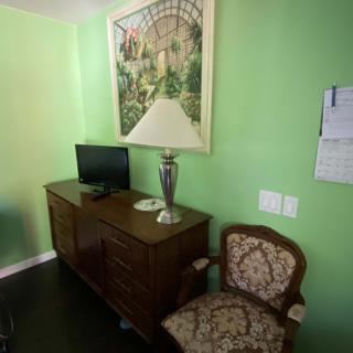 A cozy room with green walls featuring a wooden cabinet, a small television, an elegant vase, and an ornate chair beneath a large framed artwork.