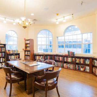 A cozy library with wooden shelves and a reading table, illuminated by natural light.