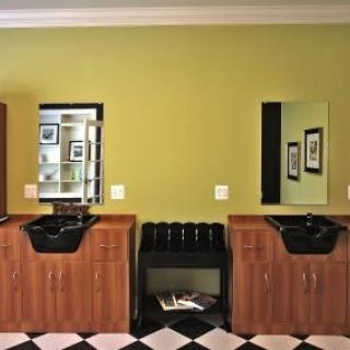 A modern barbershop interior featuring two wooden stations with black sinks and mirrors against a green wall, complemented by a black and white checkered floor.