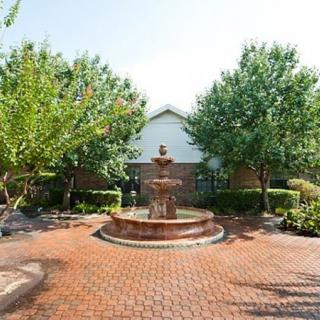 A tranquil garden setting featuring a central fountain surrounded by lush trees and a brick-paved walkway.