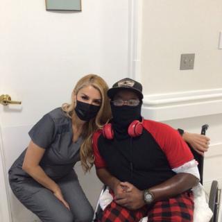 Two people in a healthcare setting wearing masks, one person is seated in a wheelchair.