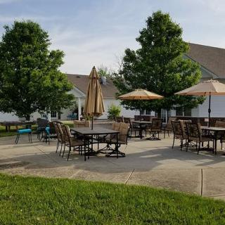 An outdoor patio area with tables, chairs, and umbrellas surrounded by greenery and a building.