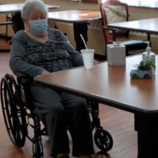 An elderly woman in a wheelchair, wearing a mask, seated at a table in a care facility.