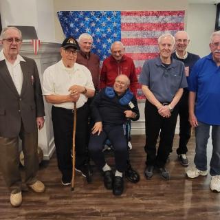 A group of elderly veterans posing together in front of an American flag indoors.