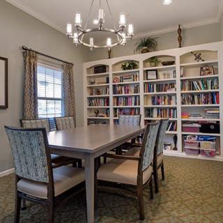 A cozy dining room with a large table, surrounded by bookshelves and illuminated by a chandelier.