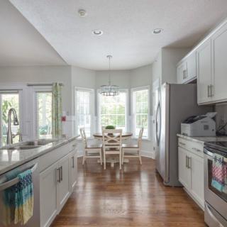 A modern kitchen with white cabinets and a dining area, featuring ample natural light and wooden flooring.