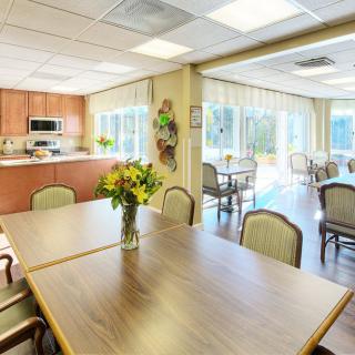 A bright and welcoming dining area with a kitchen, featuring tables, chairs, and a vase of flowers.