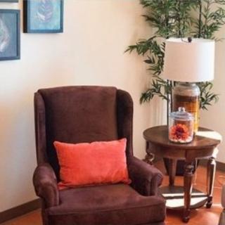A cozy living room corner with a brown armchair, a red pillow, a wooden side table with a lamp and decorative jar, and a potted plant.