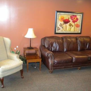 A cozy living room setup with a brown leather sofa, a white armchair, and a decorative lamp on a wooden side table. A colorful floral painting adorns the wall.
