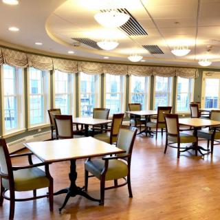 A cozy dining room with tables and chairs, featuring large windows and warm lighting.