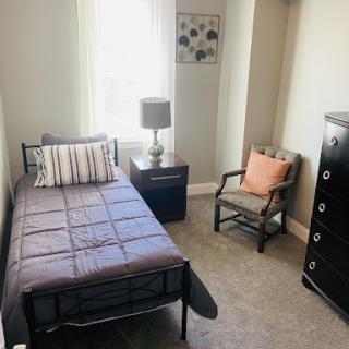 A cozy and minimalistic bedroom featuring a single bed, a nightstand with a lamp, an armchair, and a dresser.