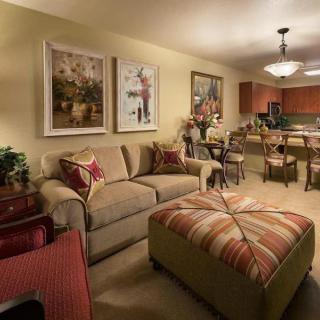 A cozy living room with a beige sofa and a large patterned ottoman, featuring warm lighting and elegant artwork. The kitchen is visible in the background.