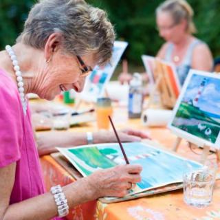 An elderly woman joyfully participates in an outdoor painting class, focusing on her watercolor artwork.