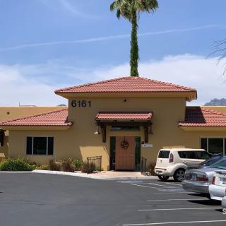A sunny day view of a building with a red-tiled roof, surrounded by parked cars and a palm tree in the background.
