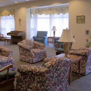A cozy living room with upholstered chairs and sofas arranged around a coffee table, illuminated by natural light from large windows.