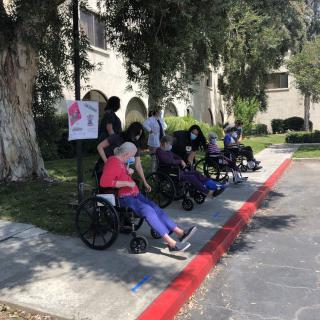 A group of elderly individuals in wheelchairs sitting on a sidewalk outside, surrounded by trees.