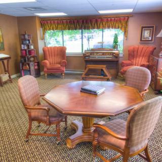 A warmly decorated conference room with a central wooden table surrounded by upholstered chairs, complete with a bookcase and large window providing natural light.