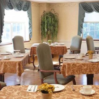 A cozy dining room with elegantly set tables and upholstered chairs, featuring decorative curtains and a potted plant.