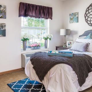 A cozy bedroom with a neatly made bed, decorative pillows, and a bedside table with a lamp. The window allows natural light to fill the room, complemented by flowers and decor on the walls.