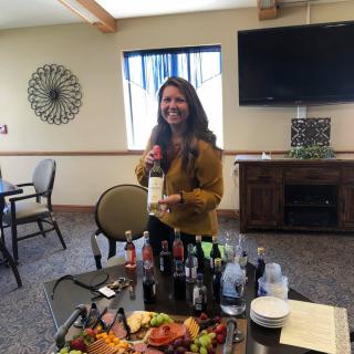 A woman holding a bottle of wine, standing by a table with various drinks and a food platter in a cozy room.