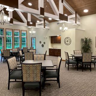 A spacious dining area with multiple tables and chairs, featuring large windows, ceiling beams, chandeliers, and a carpeted floor.