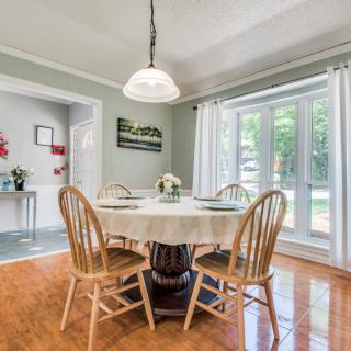 A cozy dining room featuring a round wooden table with chairs, set for a meal under a decorative chandelier, and brightened by natural light through large windows.