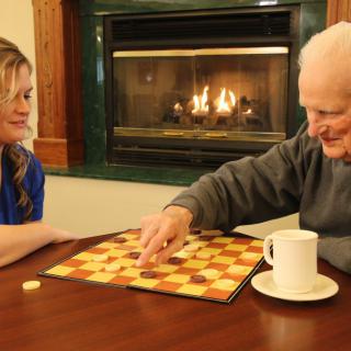 An elderly man and a woman enjoy a game of checkers by the fireplace.