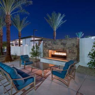 A modern outdoor patio with cozy seating and a fireplace under palm trees at dusk.
